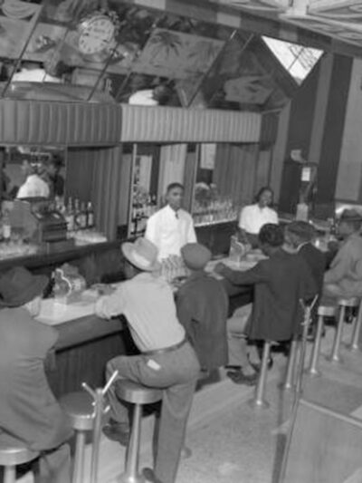 Interior photograph of one male (left) and one female bartending at the 715 Club located at 715 East 26th (twenty-sixth) Avenue in the Five Points neighborhood. A line of male customers fill the seats at the bar. They wear fedora and newsboy hats.