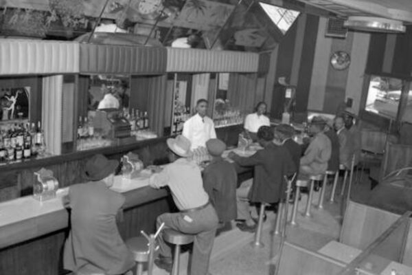 Interior photograph of one male (left) and one female bartending at the 715 Club located at 715 East 26th (twenty-sixth) Avenue in the Five Points neighborhood. A line of male customers fill the seats at the bar. They wear fedora and newsboy hats.