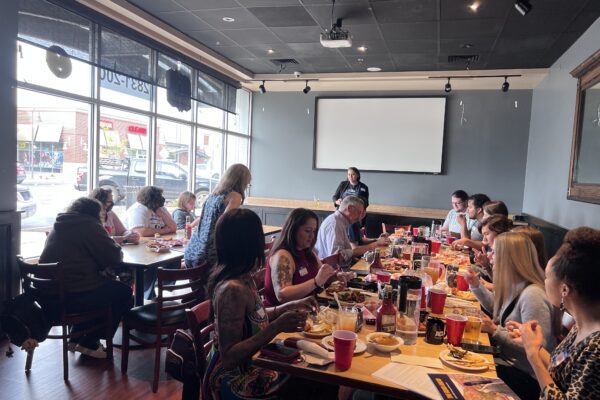 Photo of activists gathered around tables with food watching a presentation