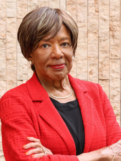 Deborah Richardson standing in front of a light brown brick wall, wearing a red jacket, arms crossed