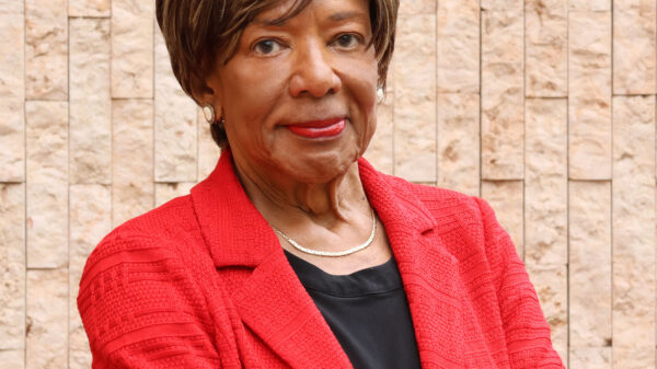 Deborah Richardson standing in front of a light brown brick wall, wearing a red jacket, arms crossed