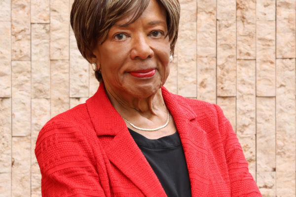 Deborah Richardson standing in front of a light brown brick wall, wearing a red jacket, arms crossed