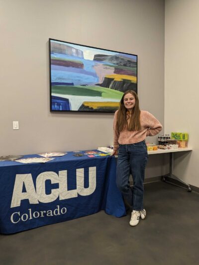 An intern stands in front of a table with a blue ACLU of Colorado tablecloth and an assortment of printed materials