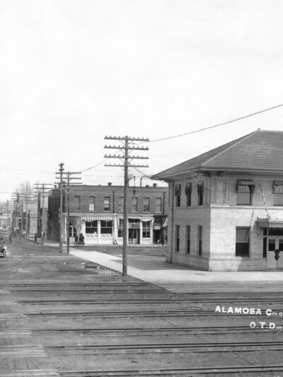 Black and white photograph of a railroad crossing with multiple rails, in a town, next to roads