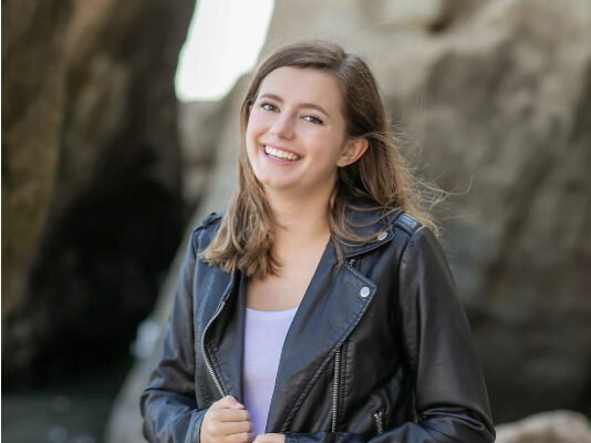 Headshot photo of Maya Walthall, smiling, standing on a beach in front of a rock formation