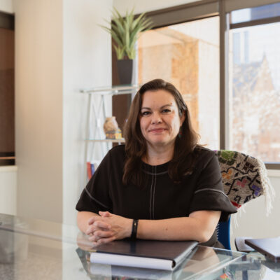 Headshot photo of Olivia Mendoza smiling, sitting at a desk, in front of a window