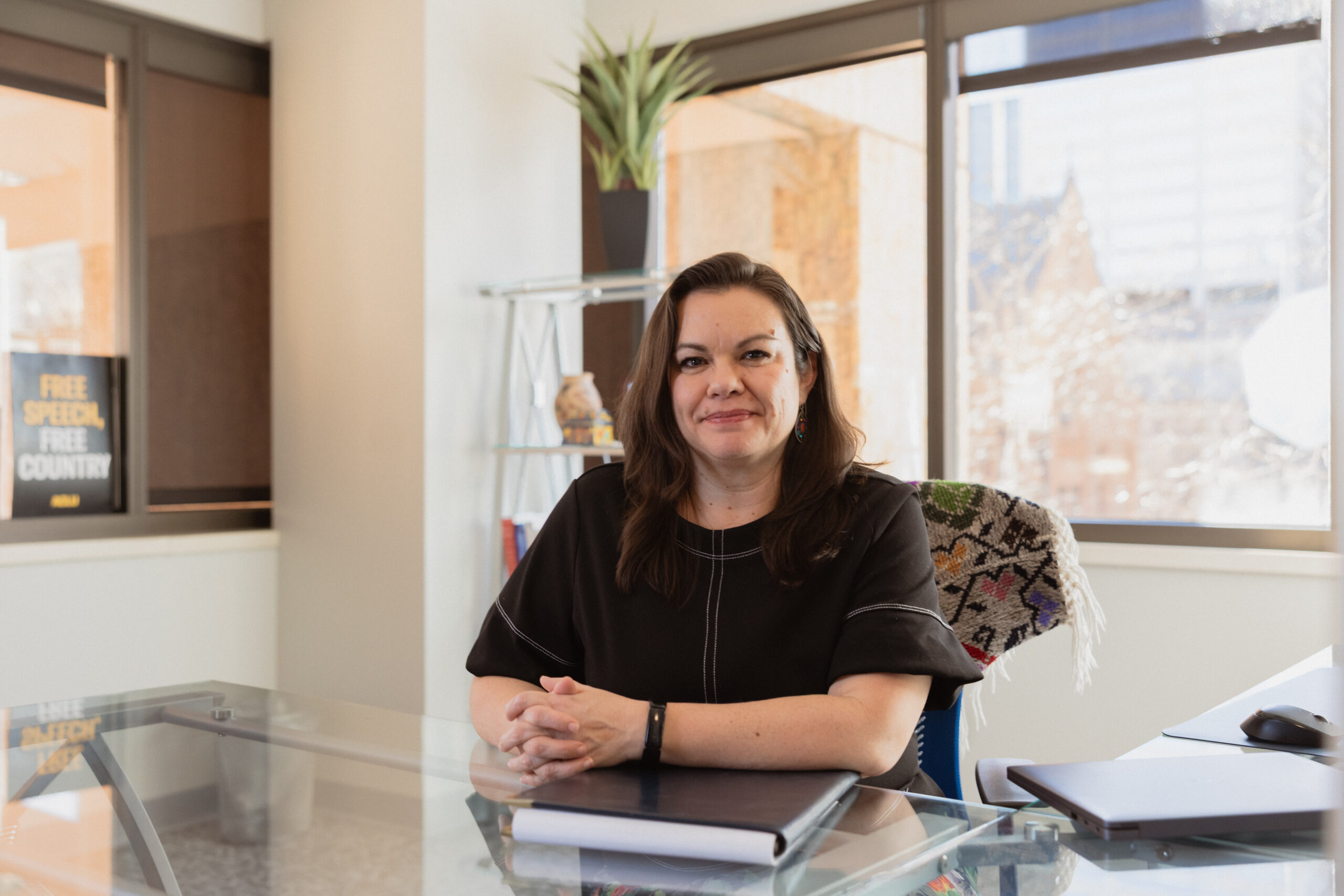 Headshot photo of Olivia Mendoza smiling, sitting at a desk, in front of a window