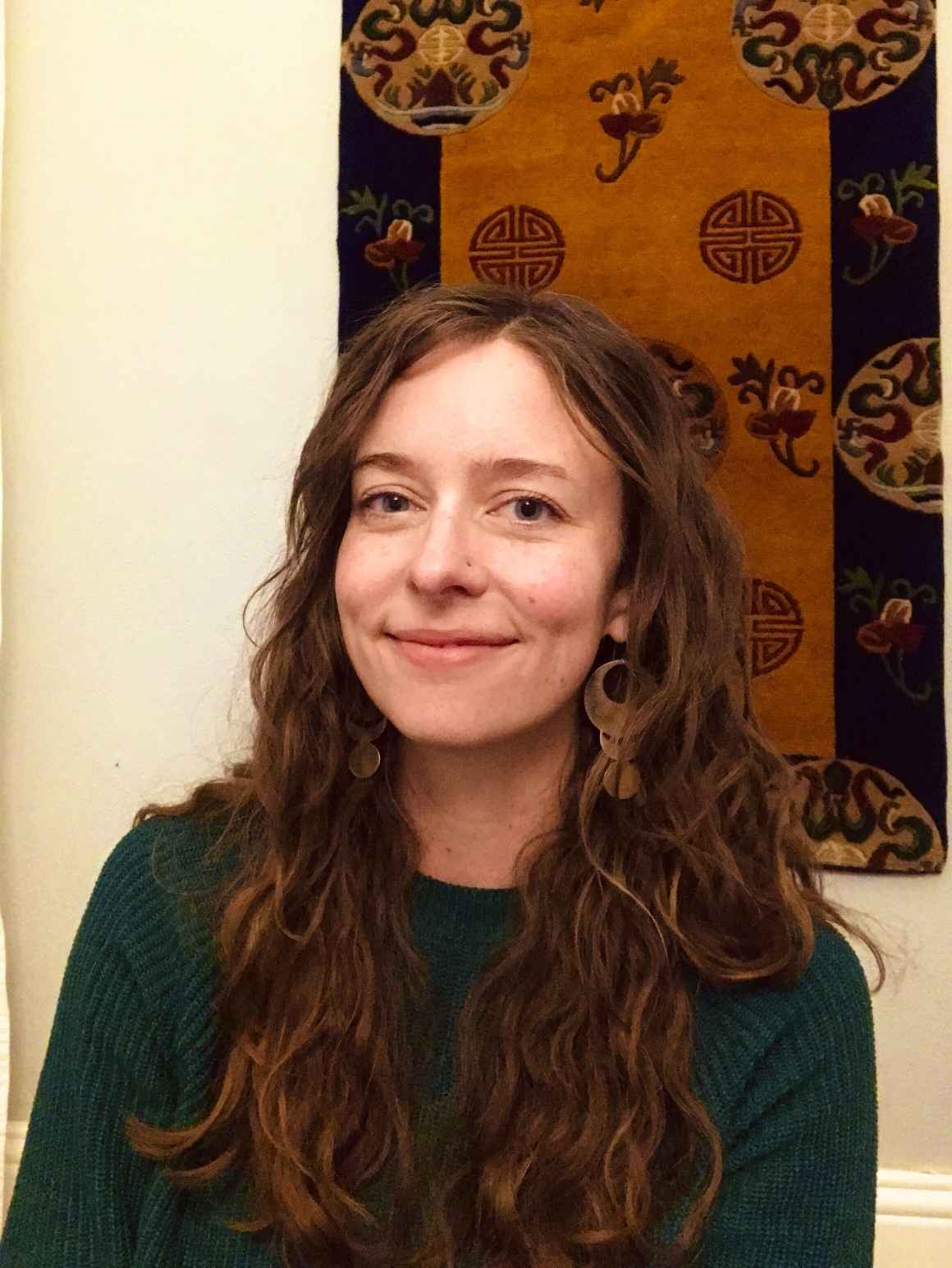 Kate Falkenhart, ACLU of Colorado operations manager, smiling, in front of light-colored wall with a brown and black tapestry hanging