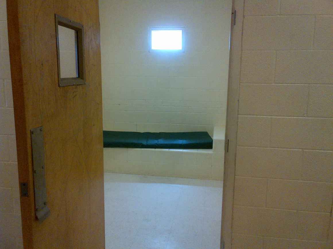 Photo of a small detention cell with white floors, walls and a mattress.