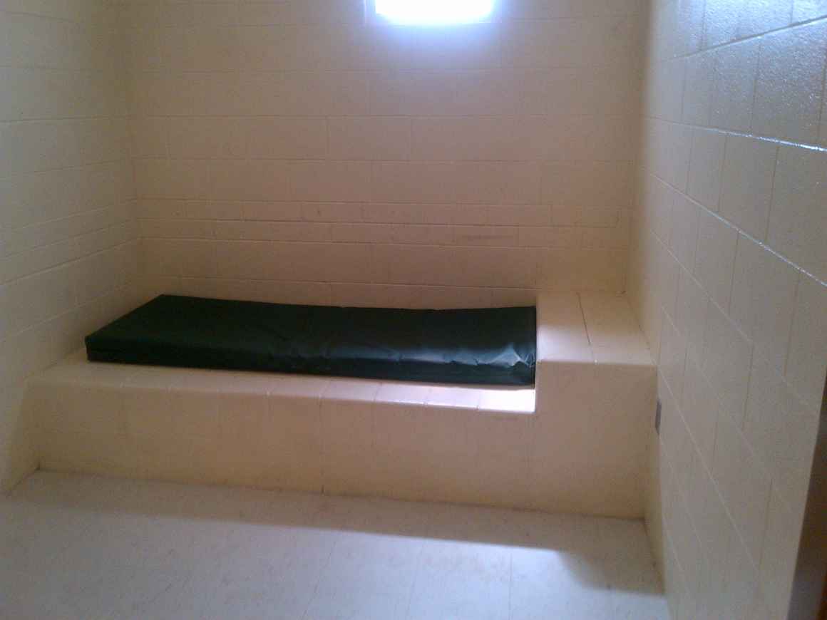 Photo of a small detention cell with white floors, walls and a mattress.