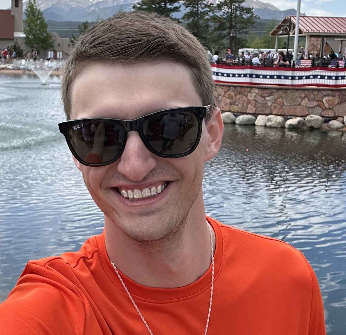 Selfie photo of Logan Ruths, smiling, wearing sunglasses in front of a pond with a flag pole on the other side of the water