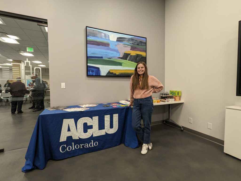 An intern stands in front of a table with a blue ACLU of Colorado tablecloth and an assortment of printed materials