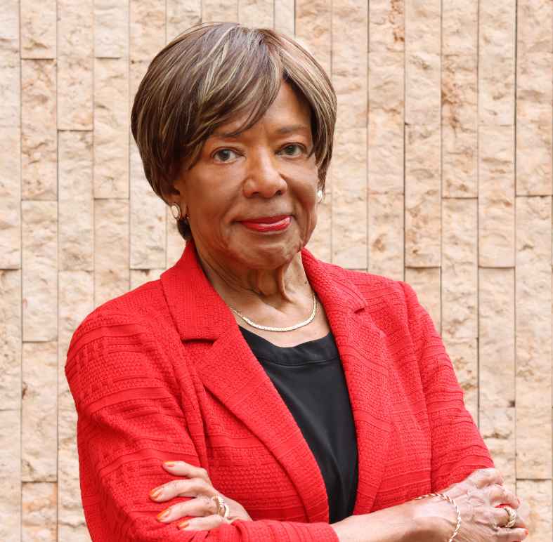 Deborah Richardson standing in front of a light brown brick wall, wearing a red jacket, arms crossed