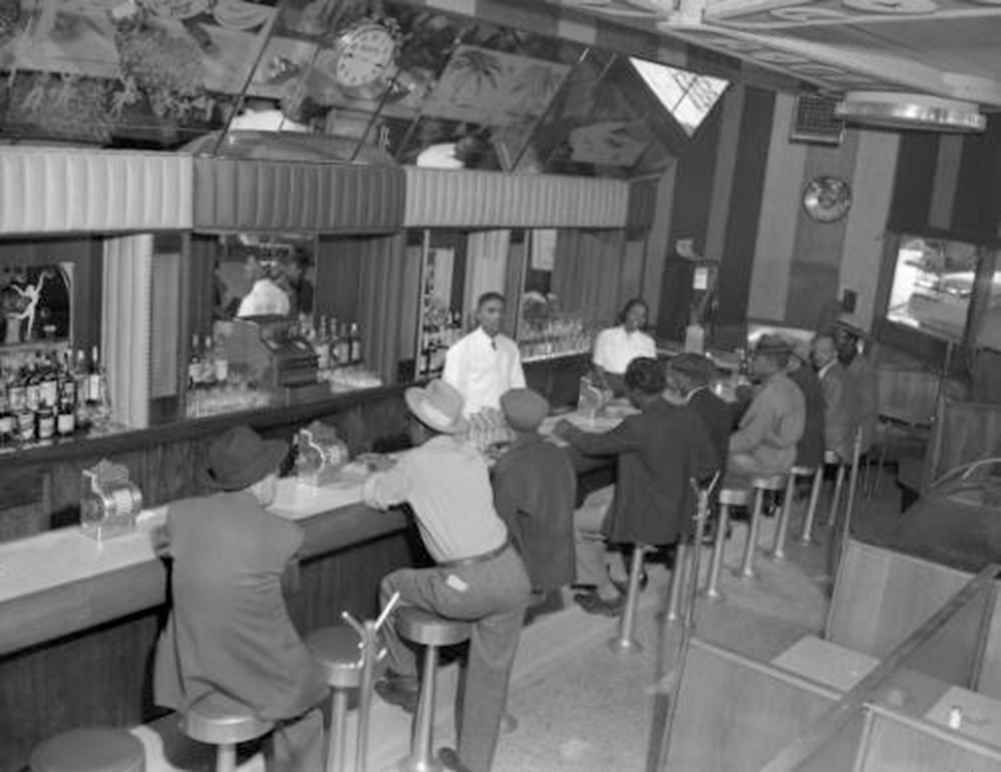 Interior photograph of one male (left) and one female bartending at the 715 Club located at 715 East 26th (twenty-sixth) Avenue in the Five Points neighborhood. A line of male customers fill the seats at the bar. They wear fedora and newsboy hats.