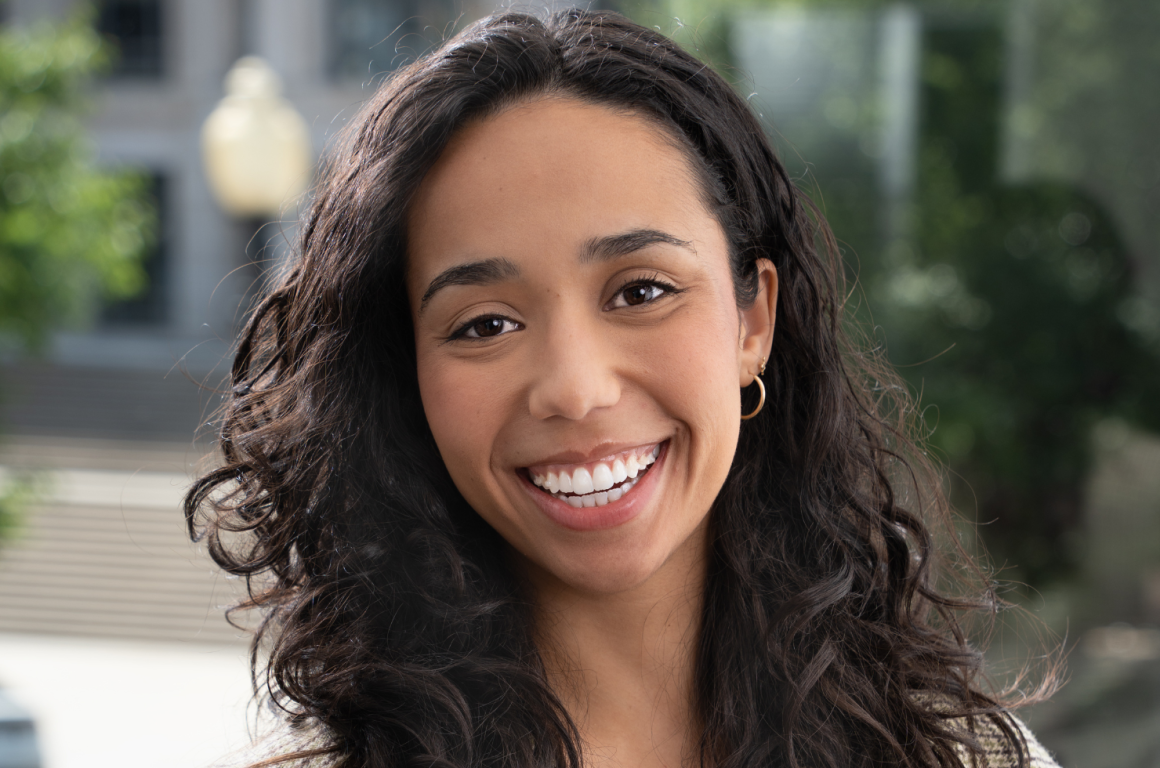 A headshot photo of Mia Bailey, smiling, with plants blurred in the background