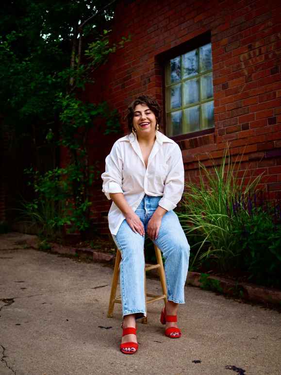 Marie Medina, smiling, outdoors, sitting on a stool in front of red brick building