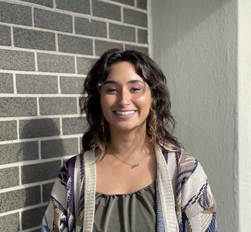 Headshot photo of Mika Alexander standing in front of a grey brick wall, smiling