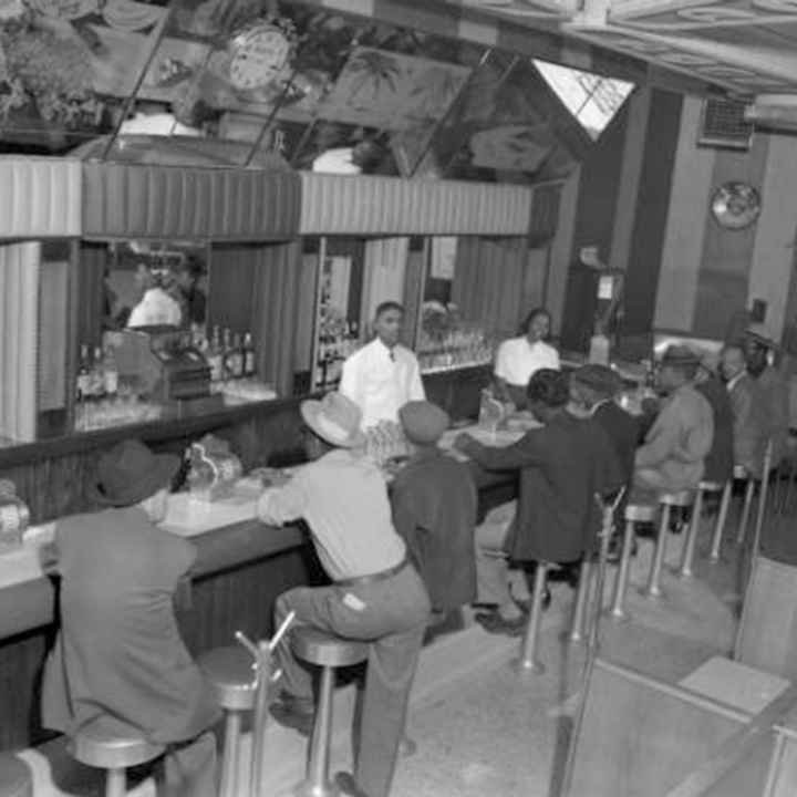 Interior photograph of one male (left) and one female bartending at the 715 Club located at 715 East 26th (twenty-sixth) Avenue in the Five Points neighborhood. A line of male customers fill the seats at the bar. They wear fedora and newsboy hats.