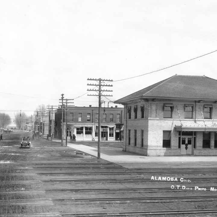 Black and white photograph of a railroad crossing with multiple rails, in a town, next to roads